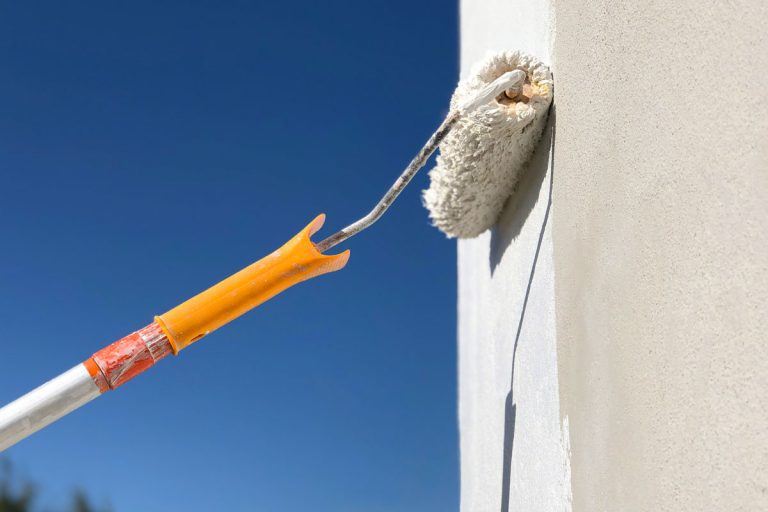 A painted using a roller to paint the exterior wall with white stucco paint, How Long Should Exterior Paint Job Last