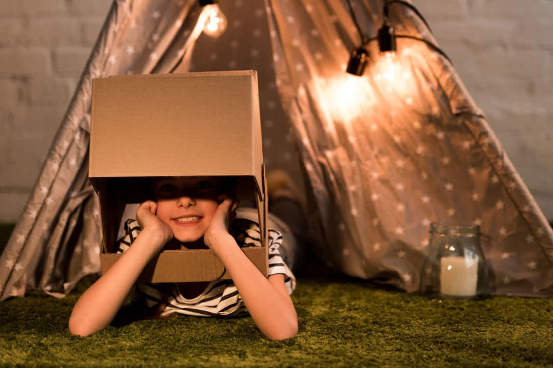A small kid putting his head in a cardboard box with a tent on the background