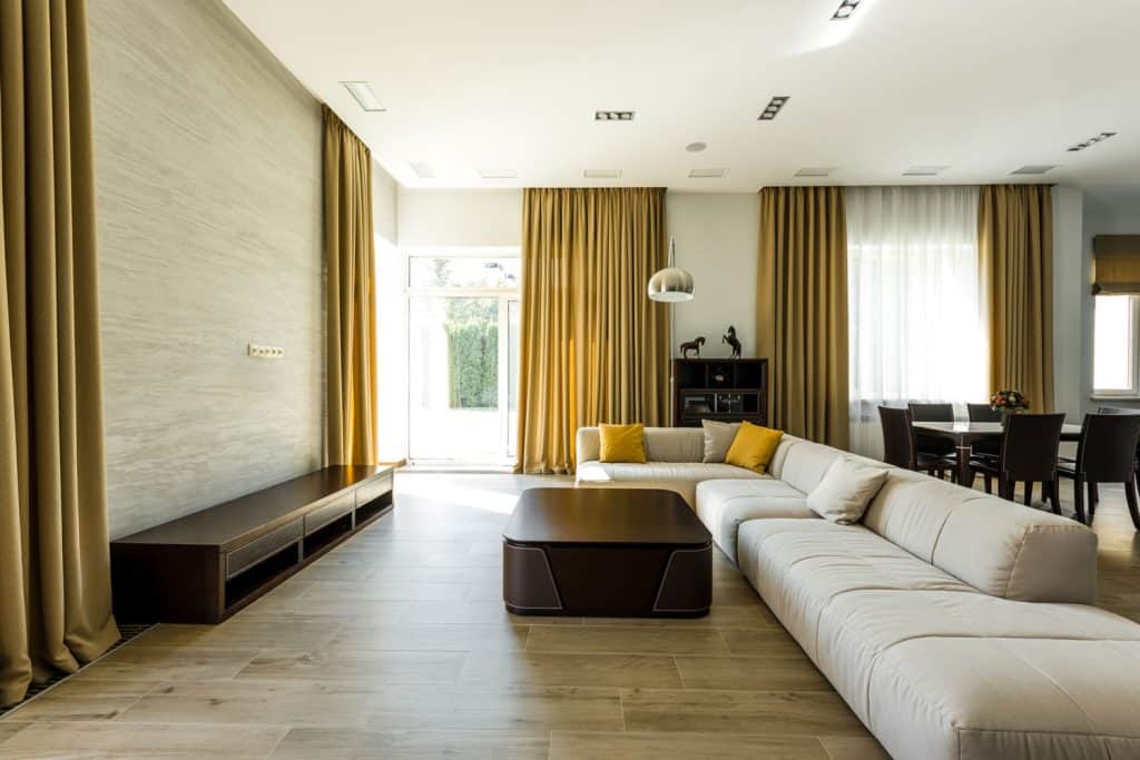 A spacious living room with a white sectional sofa, black coffee table, brown wall unit and yellow curtains