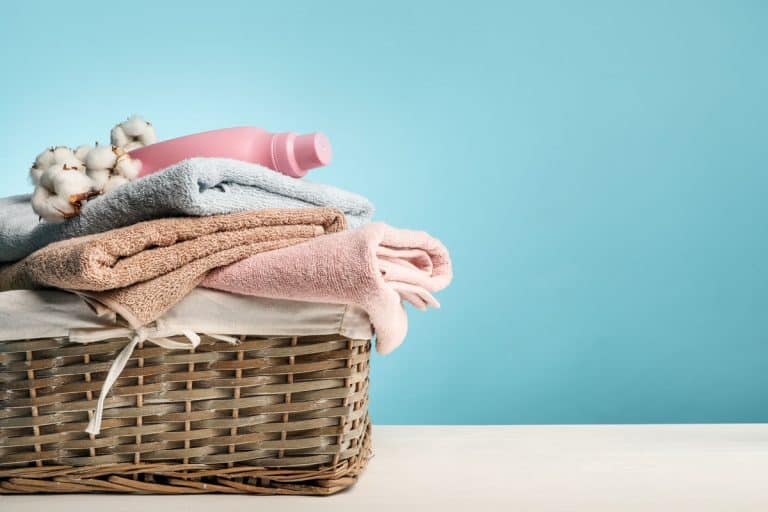 A wooden weaved laundry basket with newly cleaned laundry on a blue walled laundry room, Should You Clean A Laundry Basket (And How To)