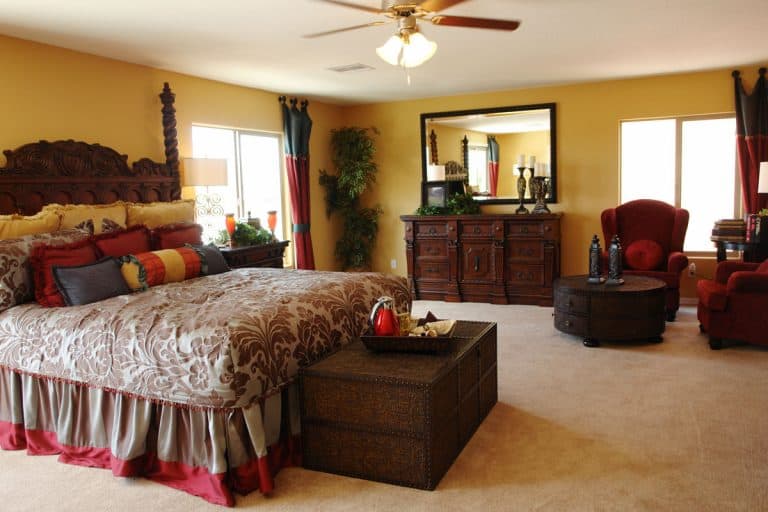 Country designed bedroom with carpeted flooring, brown furniture's, red accent chairs, and floral beddings with red throw pillows, What Color Bedding Goes With Brown Furniture?