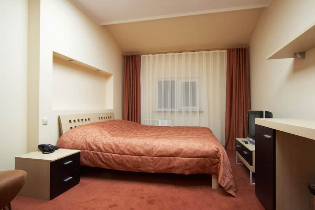 Cream painted bedroom with reddish brown carpeted floor, brown and white curtains, and a brown bedsheet