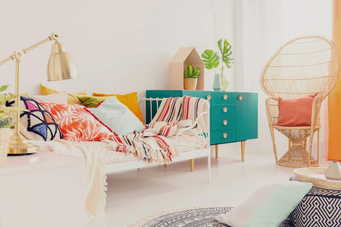 Golden lamp on nightstand in boho girl's bedroom with colorful bedding on bed, green wooden cabinet and peacock chair with pillow