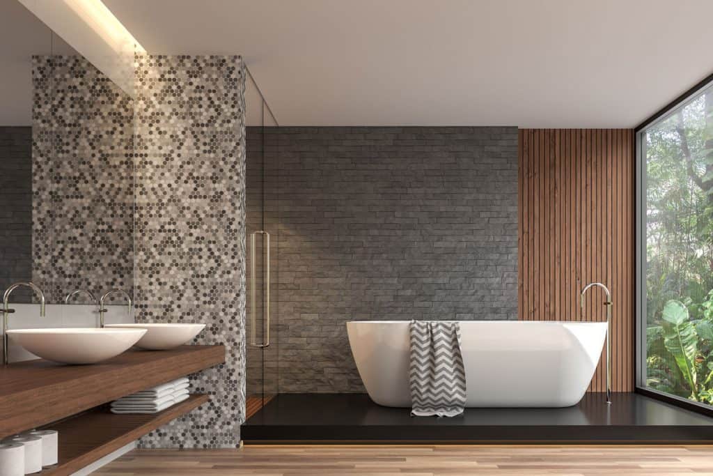 A luxurious modern contemporary bathroom with a decorative wall in the background, a raised level bathtub area, huge mirror on the vanity area, and a huge window on the right side