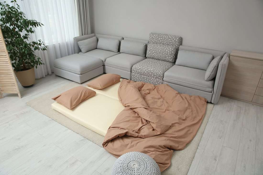 Mattress with pillows and blanket on floor near sofa. Additional sleep place for guest