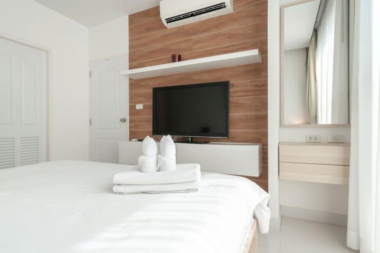 A modern contemporary bedroom with a wooden accent wall with a TV on it, a split type air-conditioning unit on top inside a white themed bed with folded towels, Where To Put TV In A Bedroom