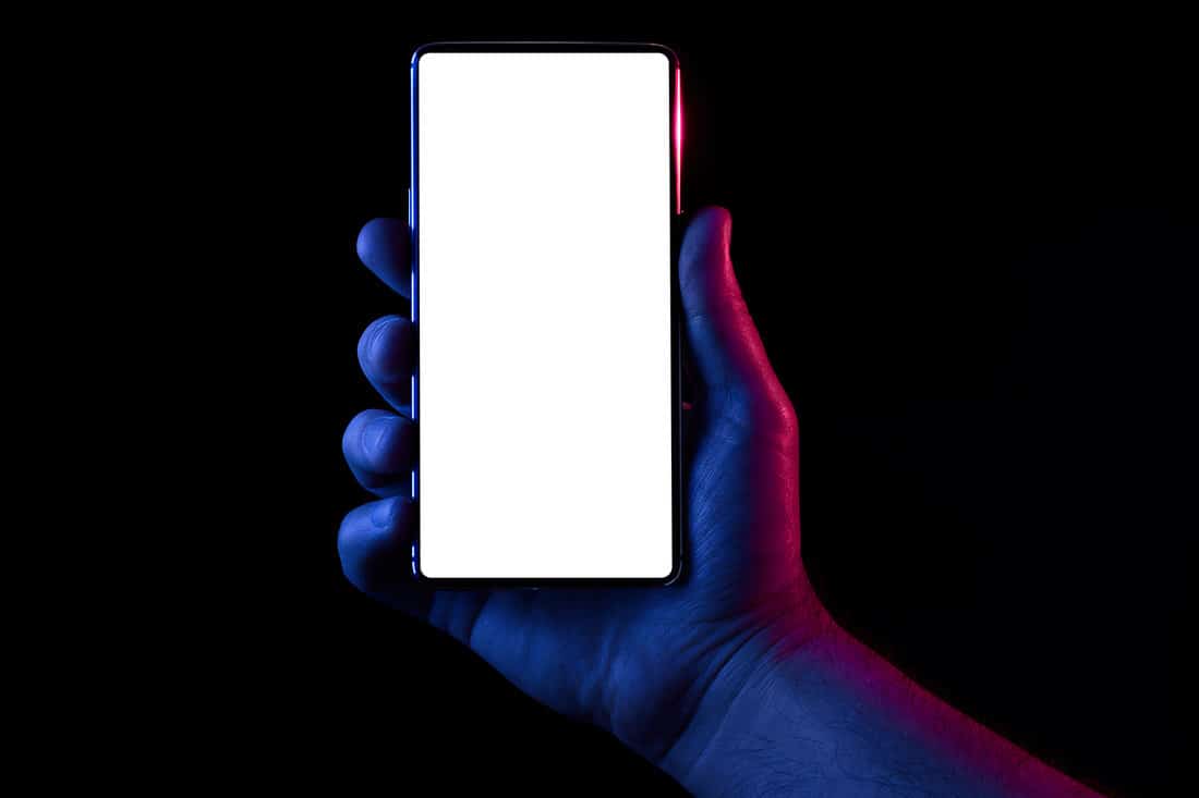 Silhouette of male hand lit with blue and red neon lights holding bezel-less smartphone on black background
