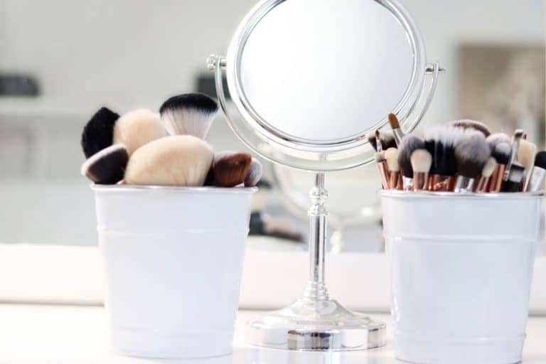 Two small white bucket containers with small makeup brushes and a round mirror, How to Organize Makeup in a Small Bathroom