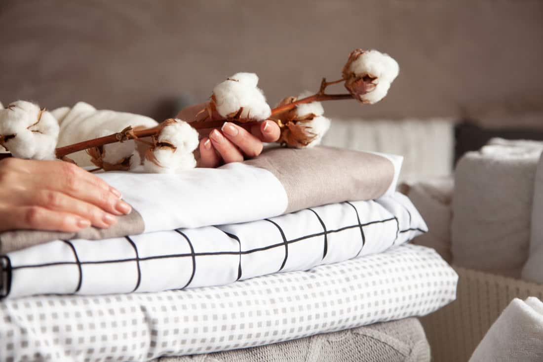 Well groomed woman hands holding the cotton branch with pile of neatly folded bed sheets, blankets and towels. Production of natural textile fiber, 4 Best Blankets For Summer [By Type of Material]