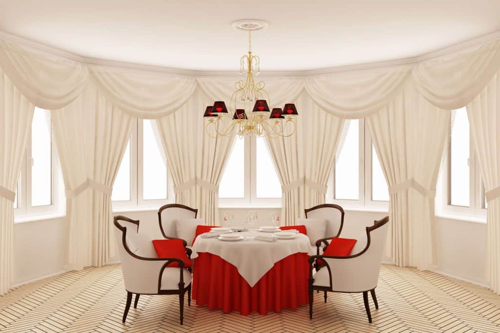 A white romantic themed dining area with a bay window with white curtains, four accent dining chairs and a red velvet table