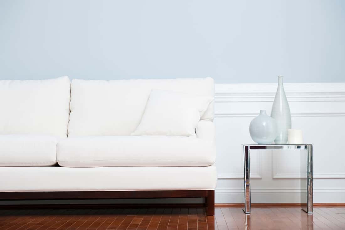 A white sofa with white throw pillows and an end table with metal framing on the side, How To Decorate Glass End Tables [5 Great Ideas!]