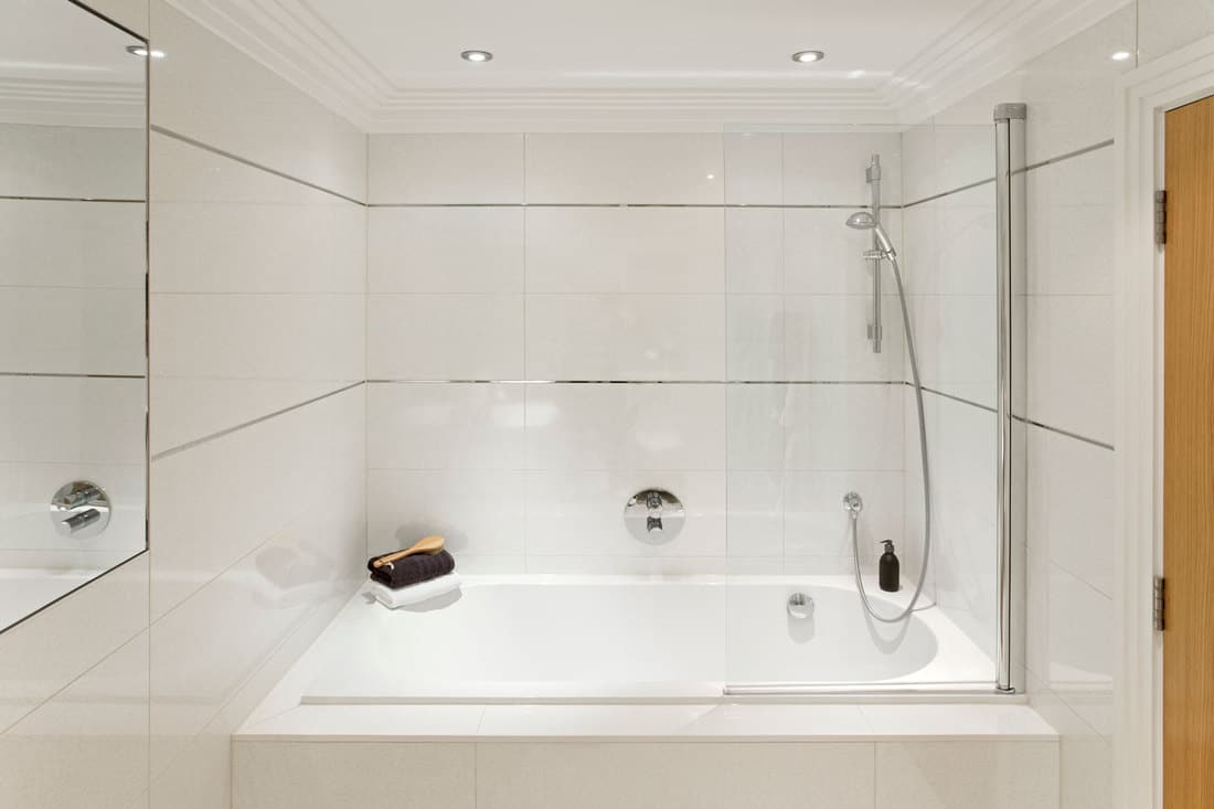 a white enamel bath with shower unit and glass shower screen in a guest's bathroom set in off-white tiles