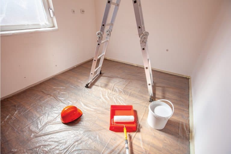 Room being prepared for painting with painters tape, plastic wrapping, paint bucket and paint roller, How Long Does It Take To Paint A Room [By Room Size]