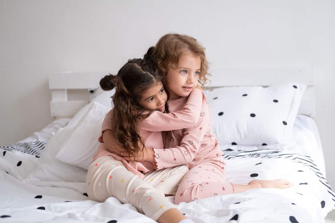 two beautiful diverse kids girls in pajamas hugging on bed in modern bright apartment. 