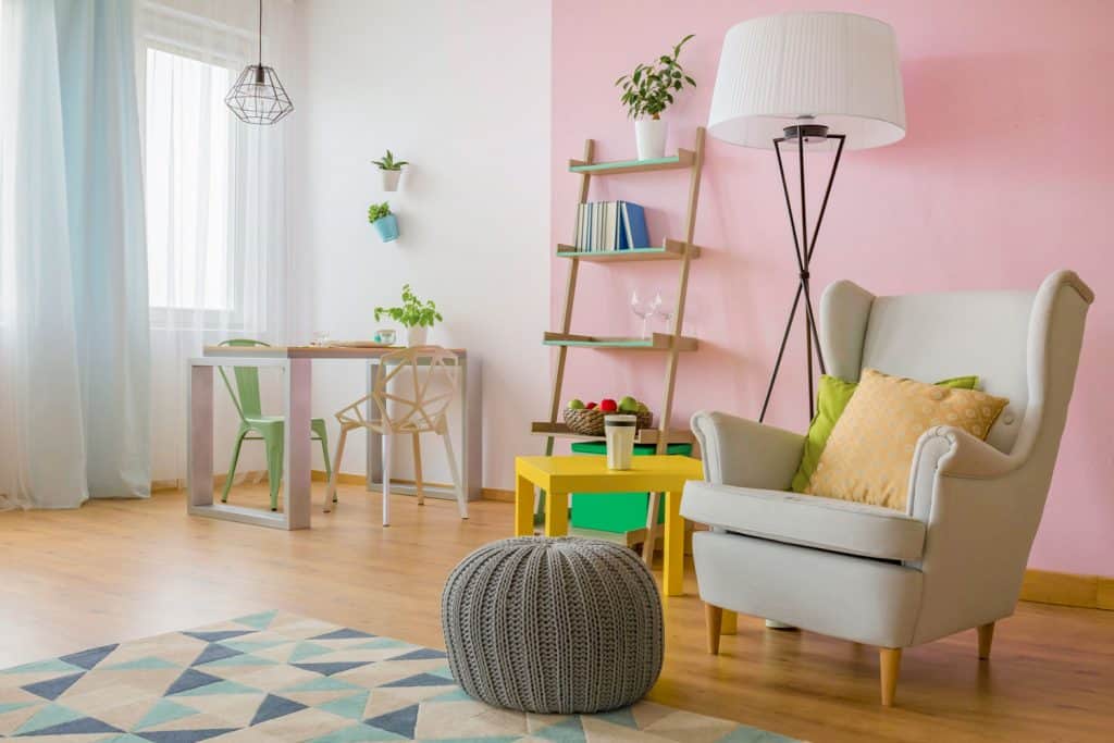 Un magnifique salon au mur rose avec des meubles blancs, un pouf gris et un abat-jour blanc