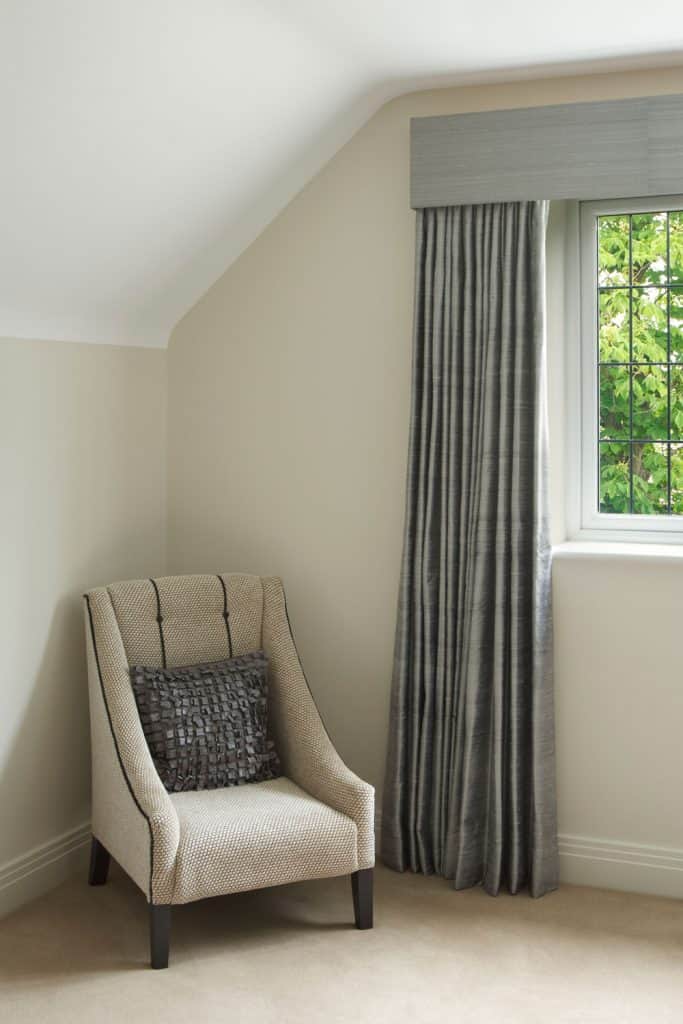 A mansard room with a small chair, beige carpet flooring and a gray curtain