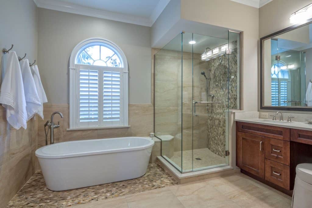 A masters bathroom with a glass wall shower with a small shower bench and a large bathtub on the side with towels hung on the wall