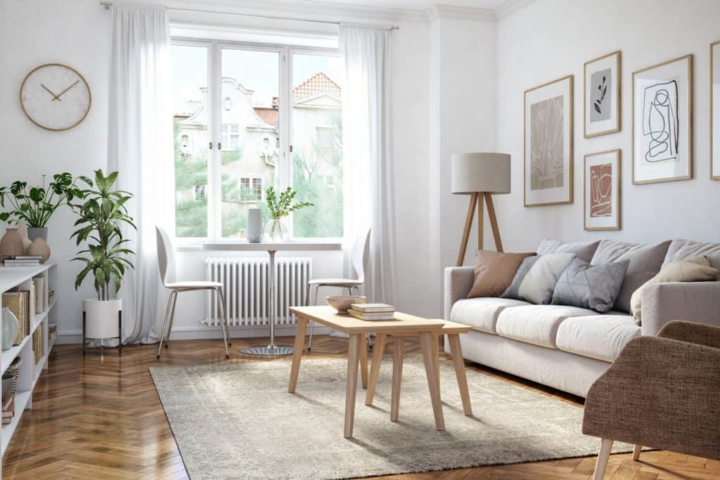 A Scandinavian themed living room with wooden flooring, beige carpet, white sofa with throw pillows and white curtains on the window