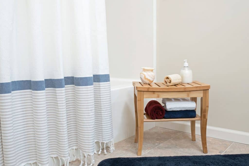 A small wooden shower bench with toiletries on the top and under placed next to a white shower curtains