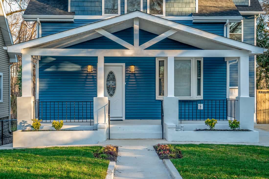Beautiful renovated craftsman style covered porch with white columns, beams, black baluster railing in front of a blue horizontal vinyl lap siding single family home in the East Coast USA.