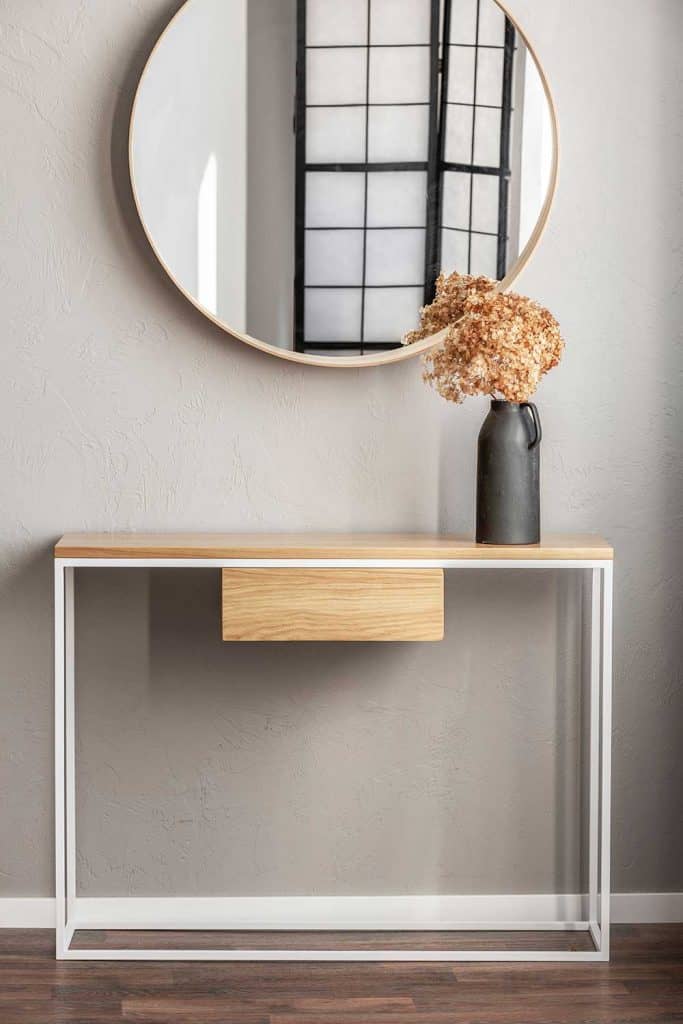 Miroir rond élégant dans un cadre en bois au-dessus d'une table console fantaisie avec des fleurs dans un vase à l'intérieur d'une chambre à la mode avec un vase beige