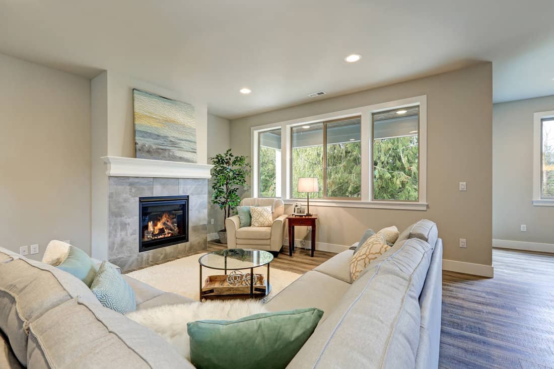 Family room interior features grey linen sectional lined with colorful pillows facing fireplace with grey tile surround across from glass top coffee table atop white soft rug.