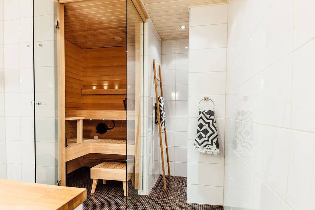 Finnish bathroom with a small wooden sauna and mosaic tile floor. Modern spa interior with glass door and Turkish towel. 