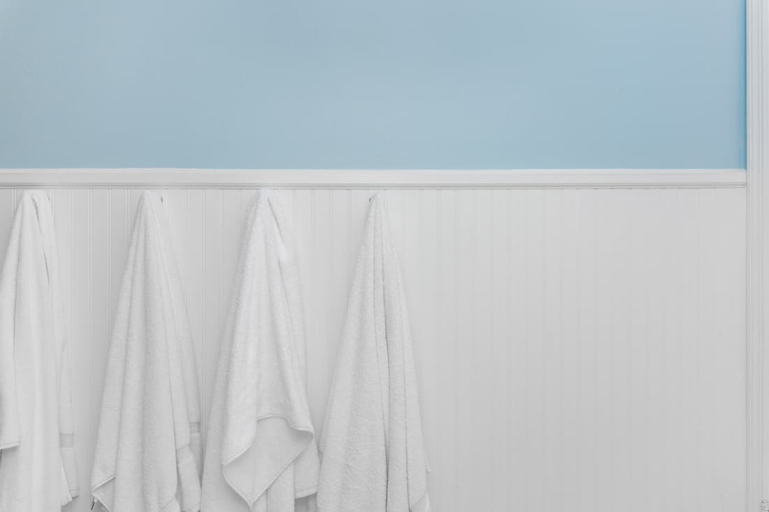 Four clean white towels hang from hooks on white beadboard or wainscoting with light blue wall paint, wood paneling wall cover for bathroom