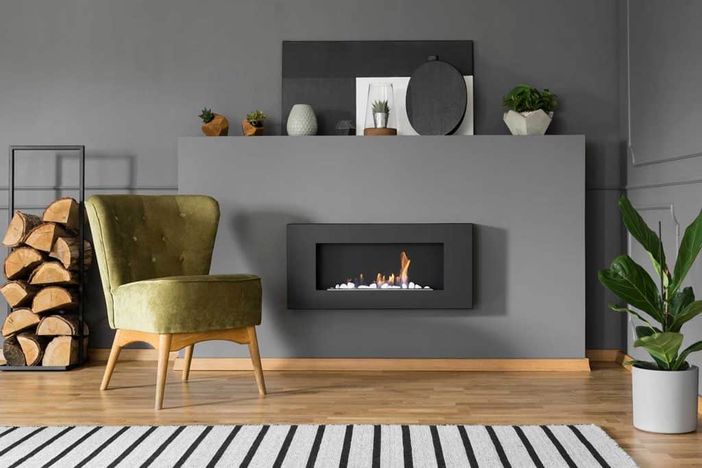 Green armchair and firewood next to fireplace in gray living room interior with plant