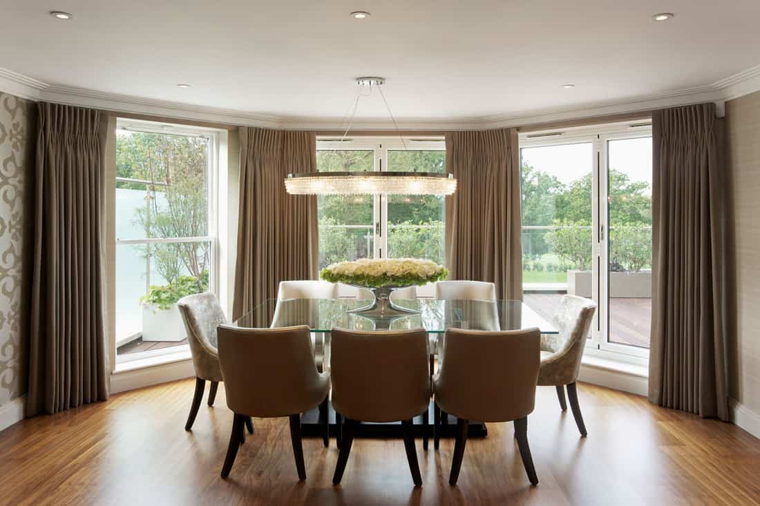 Huge bay window with brown curtains and a glass dining table with cloth seats, How Much Does A Living Room Window Cost?