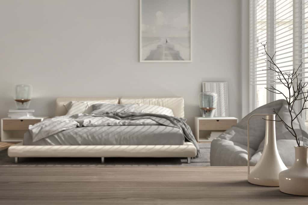 Interior of a minimalist bedroom with a king-sized bed, wooden flooring, small nightstand, and indoor plants on the side