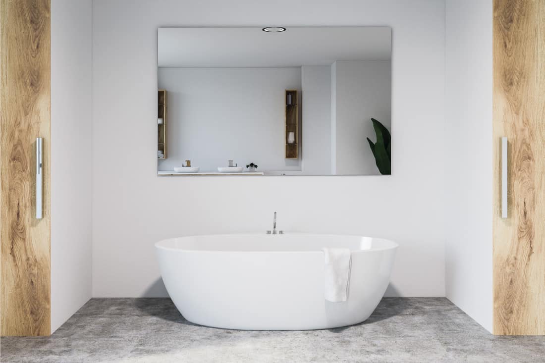 Modern bathroom interior with white walls, concrete floor, and white bathtub with large mirror hanging above it, How To Hang A Heavy Mirror [3 Ways]
