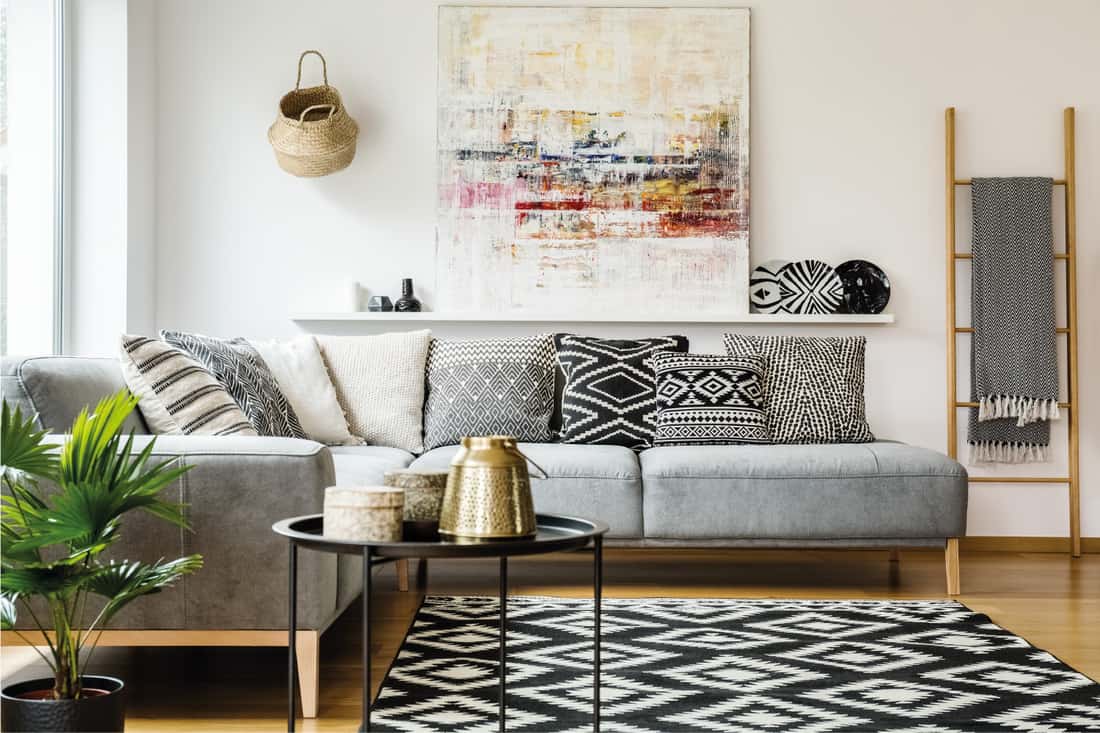 Patterned pillows on gray corner sofa in living room interior with table and painting