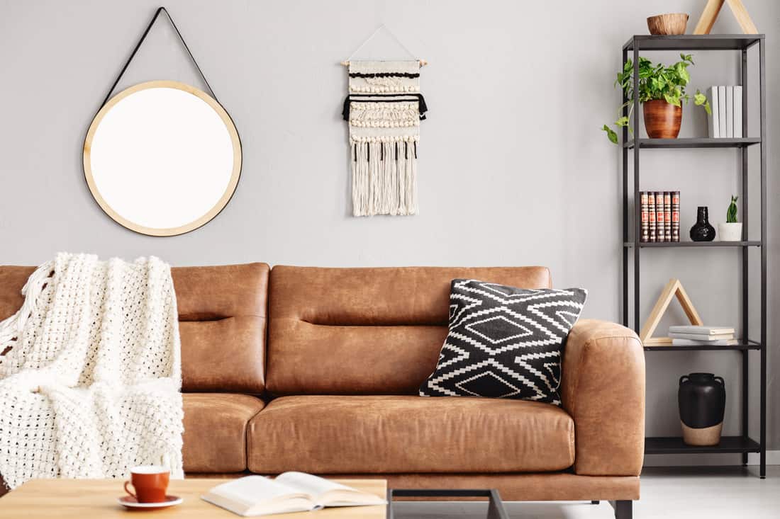 Real photo of handmade macrame and mockup poster hanging on the wall in bright sitting room interior with leather sofa and metal rack with decor and books