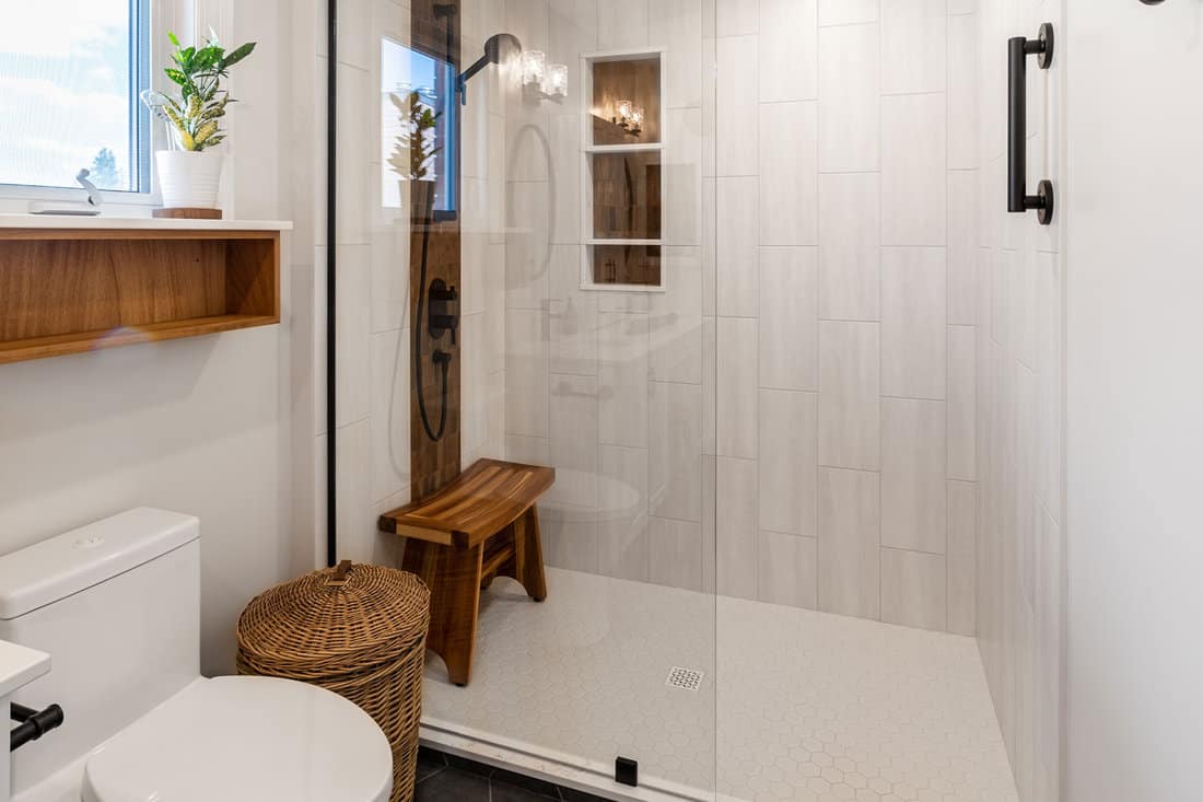 Renovated modern bathroom. Whitish tile accent wall with wood-colored tile niche. Black shower fixtures and Teak wood bench. 