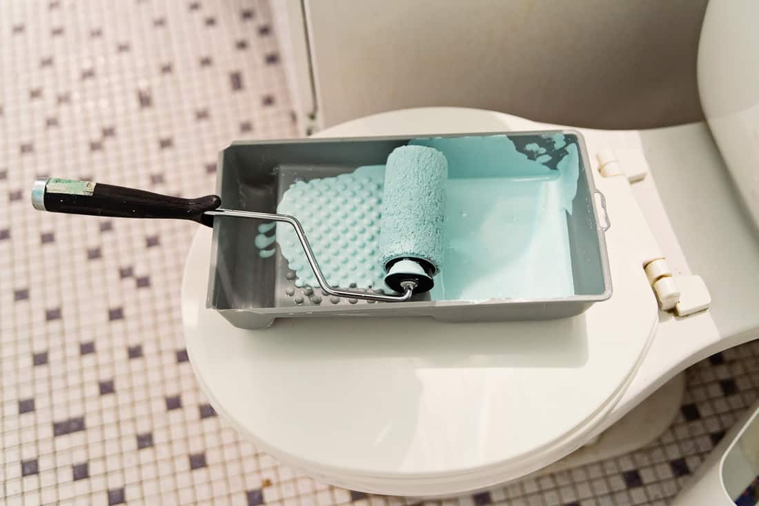 Bricolage étape par étape dans la salle de bain d'un petit appartement.  Une petite casserole de peinture bleu clair attend sur le siège des toilettes avec un rouleau recouvert de peinture. 