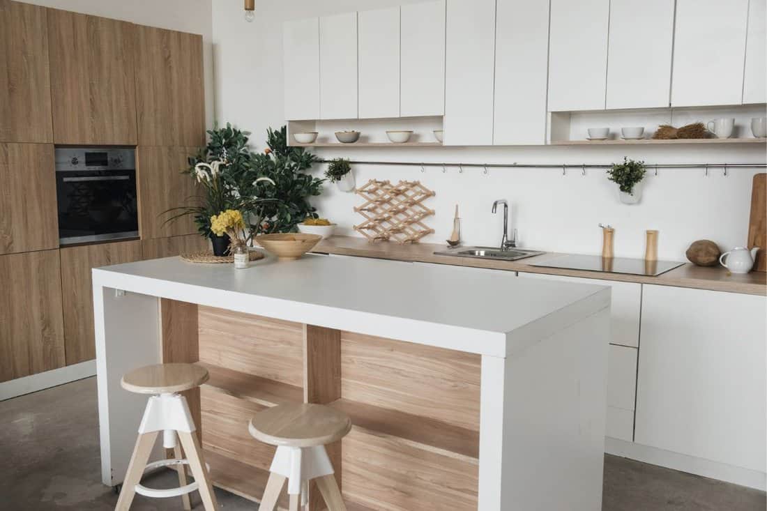 Stylish kitchen in white and brown wood. Style minimalism. Sink, table top, plants, pot, shelf for dishes. Bar stools, table.
