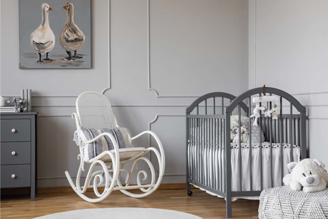 White rocking chair with pillow next to wooden cradle in elegant baby room with duck poster on the wall