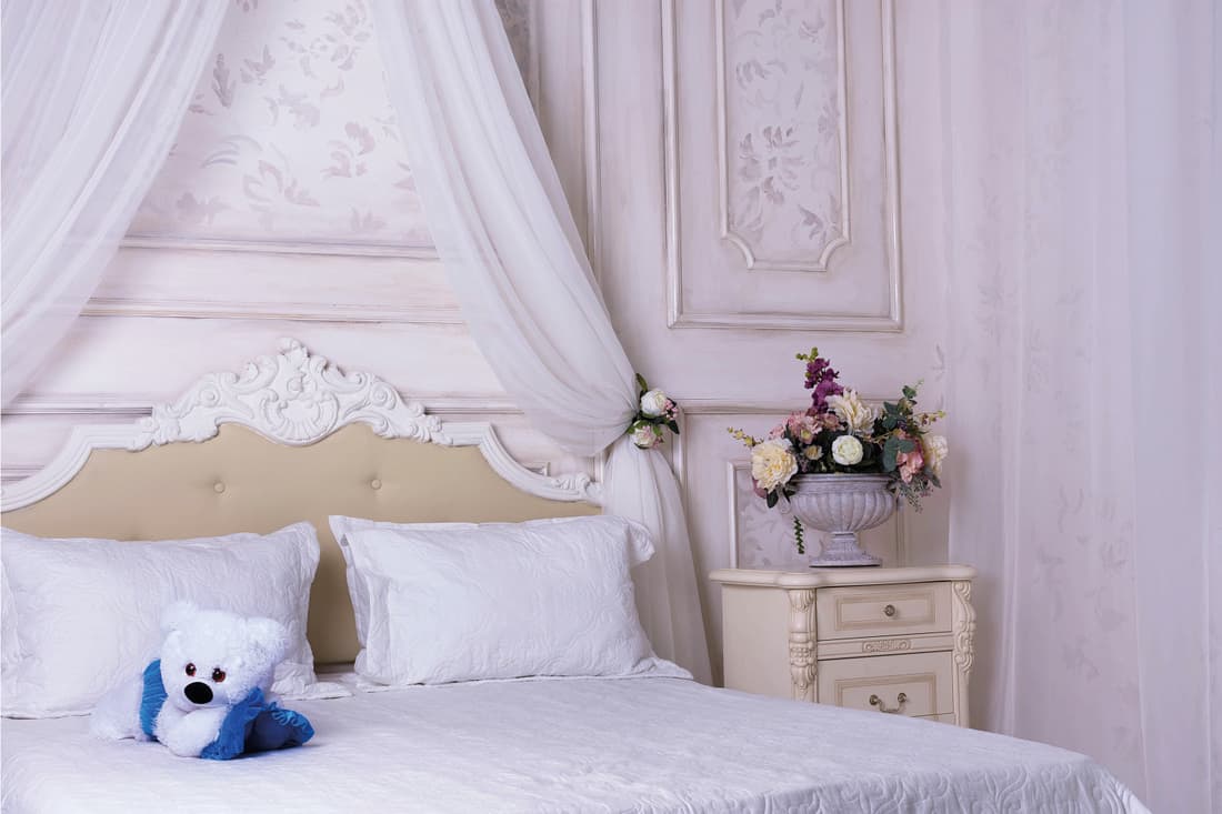 Light purple wall, nightstand, pillow, beddings, flower in the vase in the nightstand and stuffed toy on the bed