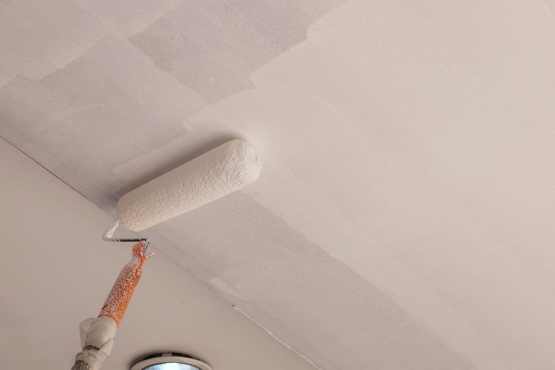 A close-up of a long-handled roller brush paints a white primer on a gypsum ceiling in a renovated home