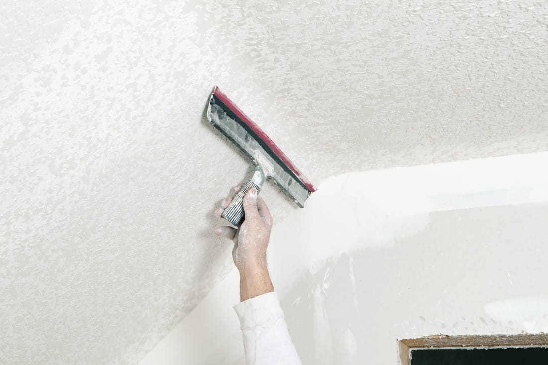 A drywall worker is troweling over sprayed drywall mud, with a rubber blade, to create a ceiling knockdown texture
