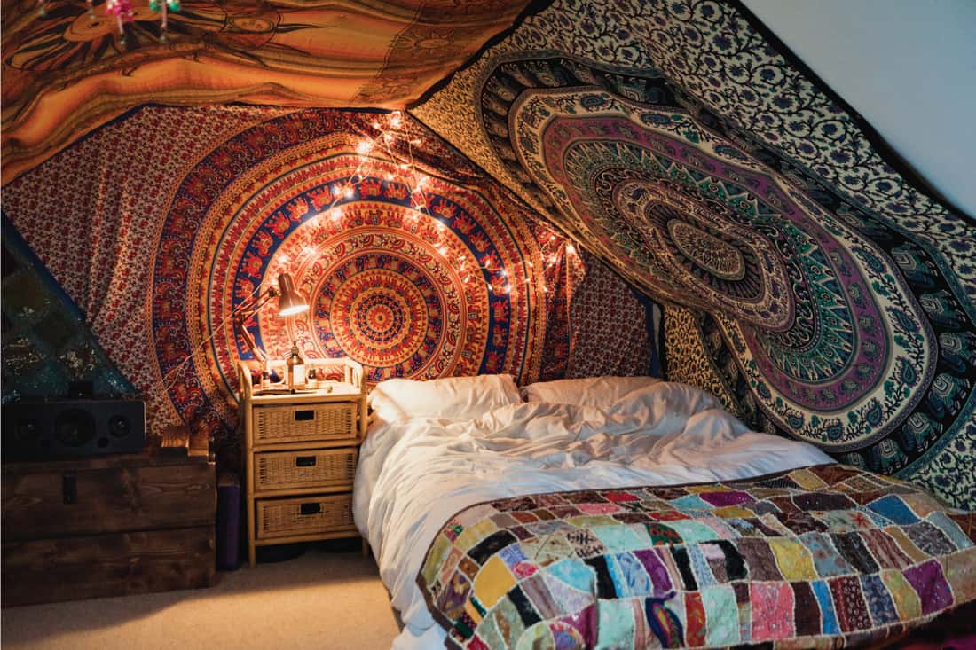 A front-view shot of a stylish bedroom interior, tapestry patterned fabrics can be seen hanging on the walls with string lights.