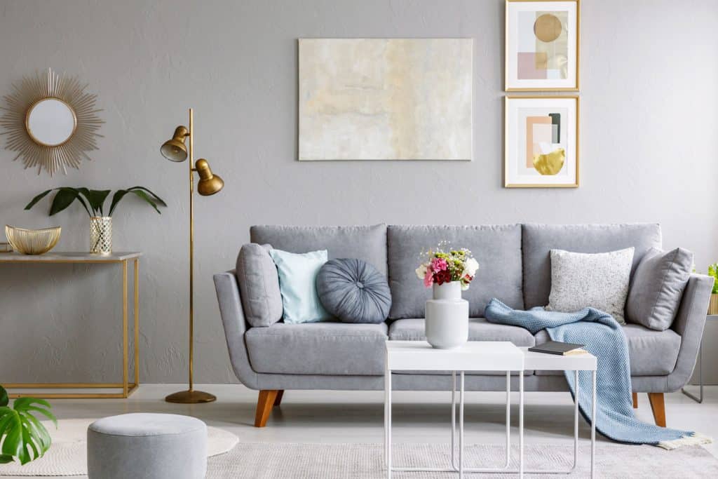A gray sofa inside a gray living room with picture frames on the wall, gray ottoman, and a golden colored floor lamp