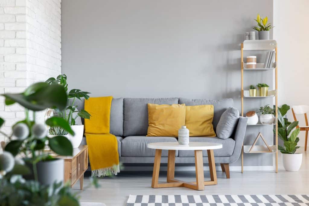 A living room with a gray sectional sofa with yellow throw pillows, a round white coffee table, and indoor plants all over the room