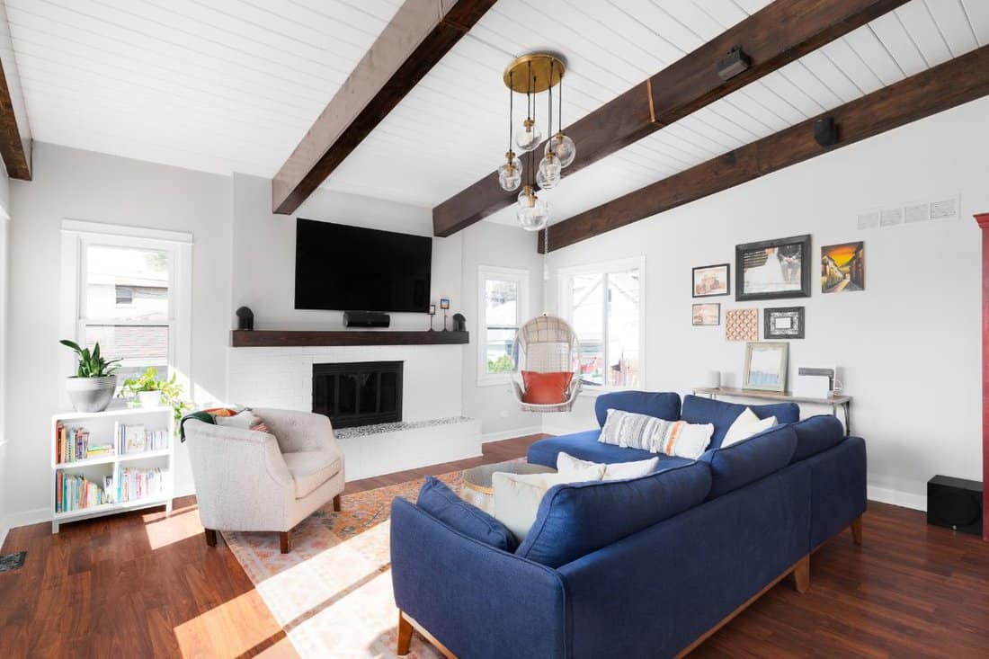 A renovated modern farmhouse living room with a blue couch, white fireplace, wood beams on a white shiplap ceiling, and a television mounted above the fireplace.