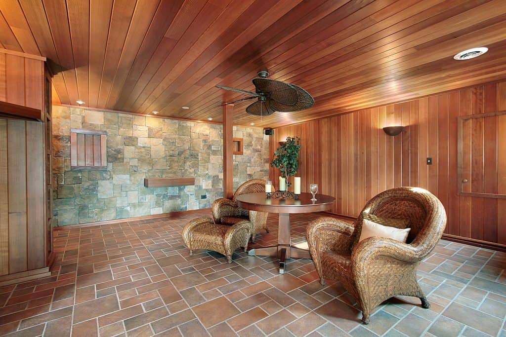 A rustic themed basement with wooden paneled walls, clay tiled flooring, and accent design wicker chairs