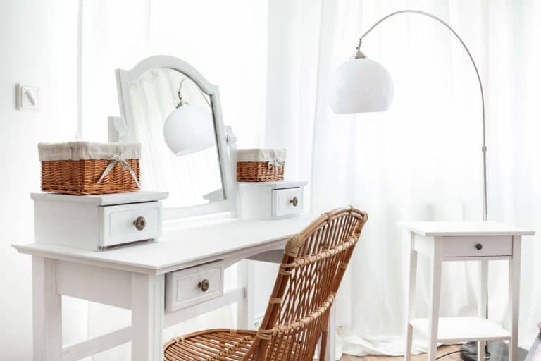 A white dresser table with small basket containers, a white framed glass mirror, and a wicker chair inside a white bedroom, How Much Does A Dressing Table Weigh?
