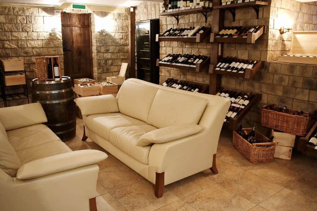 A winery rack filled with different assorted wines, two whtie loveseat sofas, and decorative stone walls