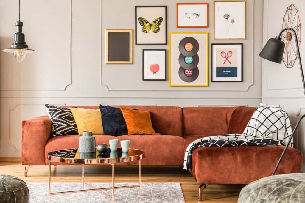 An 80's themed living room with an energizing theme of an orange velvet sofa, picture frames on the wall, and a round brass coffee table