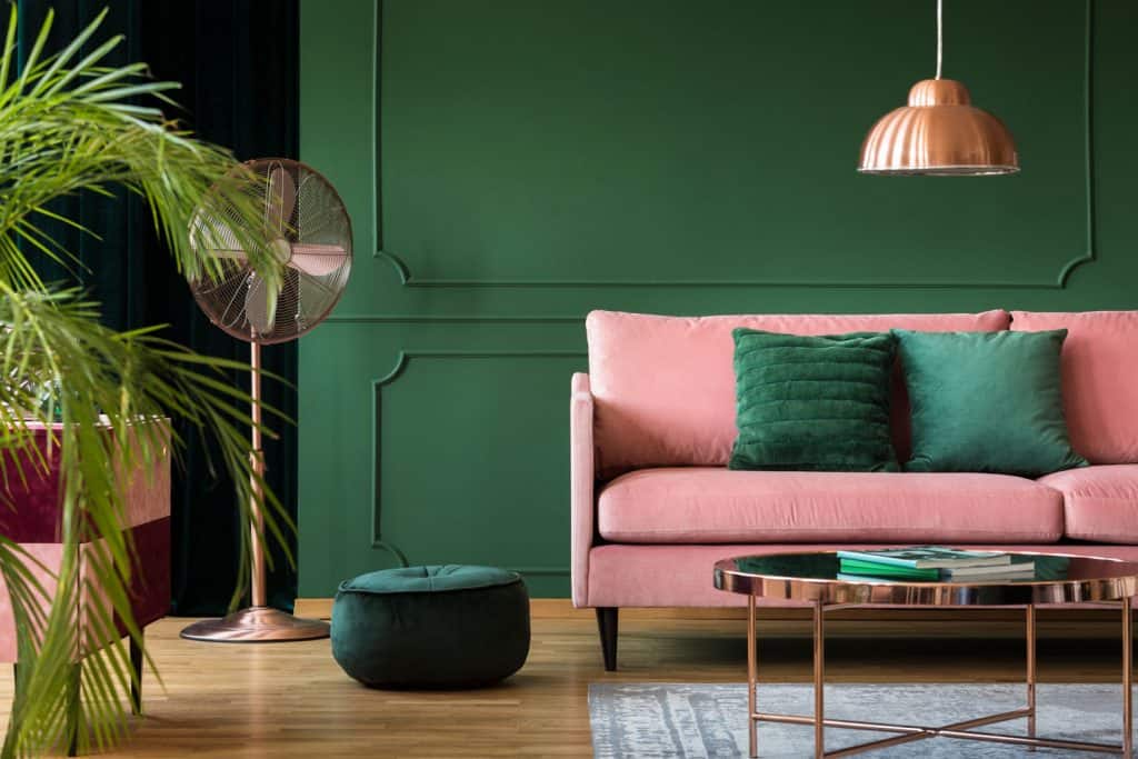 An eclectic living room with a pink sofa with green throw pillows, green wall with a brass stand fan, and a small green ottoman on the side of the sofa